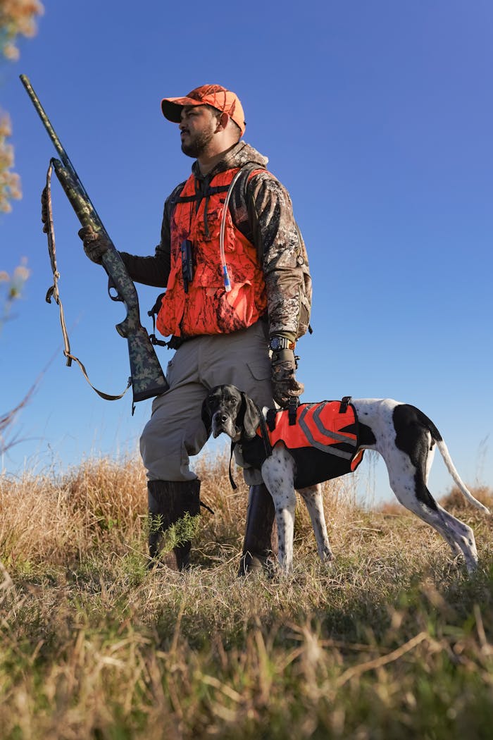 A man in hunting gear with a gun and dog standing in a grassy field under clear blue sky.