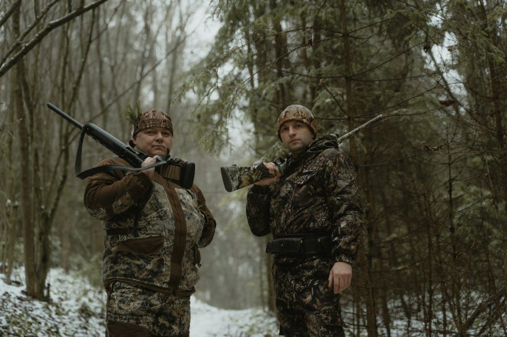 Two hunters in camouflage with rifles in a snowy forest landscape.